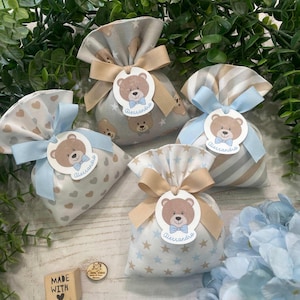 May include: Five small fabric gift bags with teddy bear tags and ribbons. The bags feature patterns of hearts, stars, and teddy bears. Each tag says "Alessandro". A wooden block with "MADE WITH" is also visible.