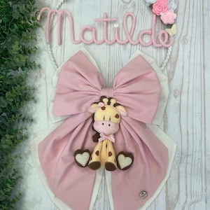 May include: A pink and white decorative wall hanging with the name "Matilde" in pink script. The design includes a large pink bow, a giraffe figure, and floral accents. The text "Sveglia, annunciando la nascita della sorellina" is above the name.