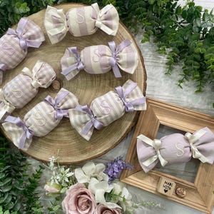 Puede incluir: Varios adornos de tela con forma de caramelo con rayas o patrones de cuadros vichy en color violeta claro y blanco. Cada adorno está atado con cintas de satén en ambos extremos y bordado con la letra S. Un adorno descansa dentro de un marco de madera.