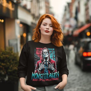 May include: Black sweatshirt featuring a graphic of Frankenstein with the text "MONSTER PARTY" and "BOO-TIFUL NIGHTMARE" in blue and teal. The design includes red and blue accents. The person in the image is wearing gray pants.