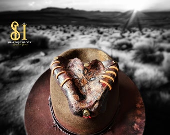 May include: A brown cowboy hat with a detailed leather snake design on the front. The snake design features intricate carvings and stitching, with a bullet casing and red string detail. The hat has a wide brim and a decorative band.