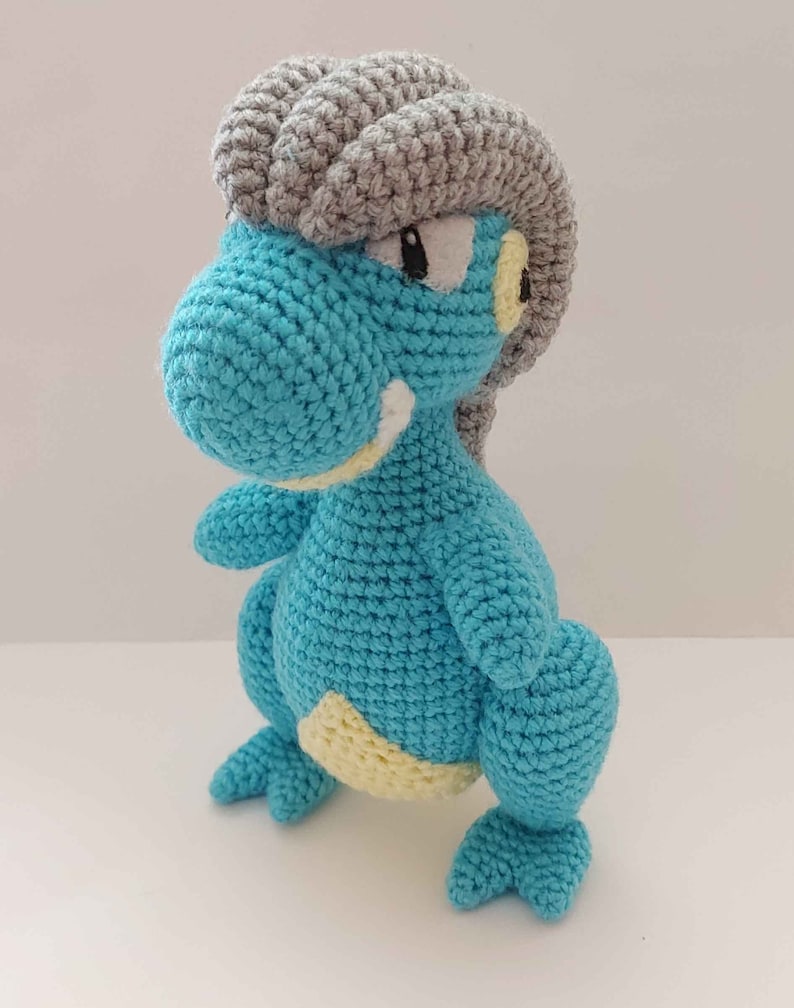 May include: A crocheted blue and gray dinosaur toy with yellow accents. The dinosaur has a spiky gray crest on its head.