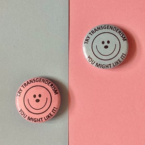 May include: Two round buttons with a smiley face and the text "Try Transgenderism You Might Like It!" on a pink and blue background.