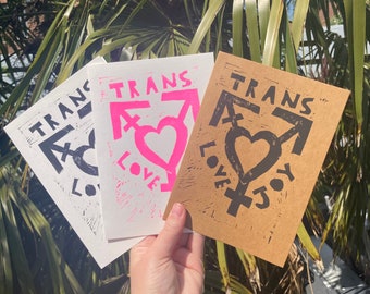 May include: Three cards with the text "Trans Love" printed on them. The cards are made of different materials, one is white, one is pink, and one is brown. The text is in black ink on the white and brown cards, and in pink ink on the pink card. The cards feature a stylized symbol of a heart with a transgender symbol.