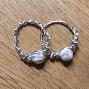 Peut inclure: Deux bagues en fil d'argent. Chaque bague est ornée d'une perle blanche et grise marbrée. Le fil est enroulé de manière complexe autour de la base et de la perle de la bague, créant un aspect texturé. Les bagues sont présentées sur une surface en bois.