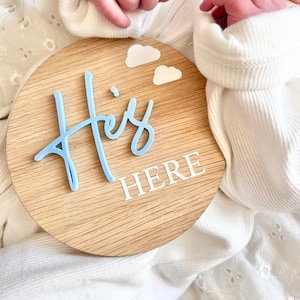 May include: A light blue wooden round sign with the words "He's Here" in white lettering. There are two white clouds on the sign. A baby is holding the sign.