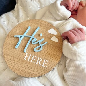 May include: A light blue wooden circle sign with the words "He's Here" in white lettering. Two small white clouds are above the word "He's".