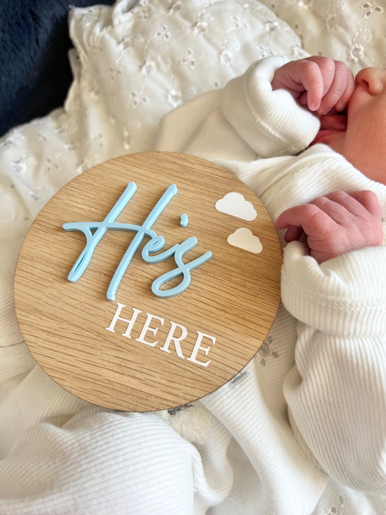 May include: A light blue wooden circle sign with the words "He's Here" in white lettering. Two small white clouds are above the word "He's".
