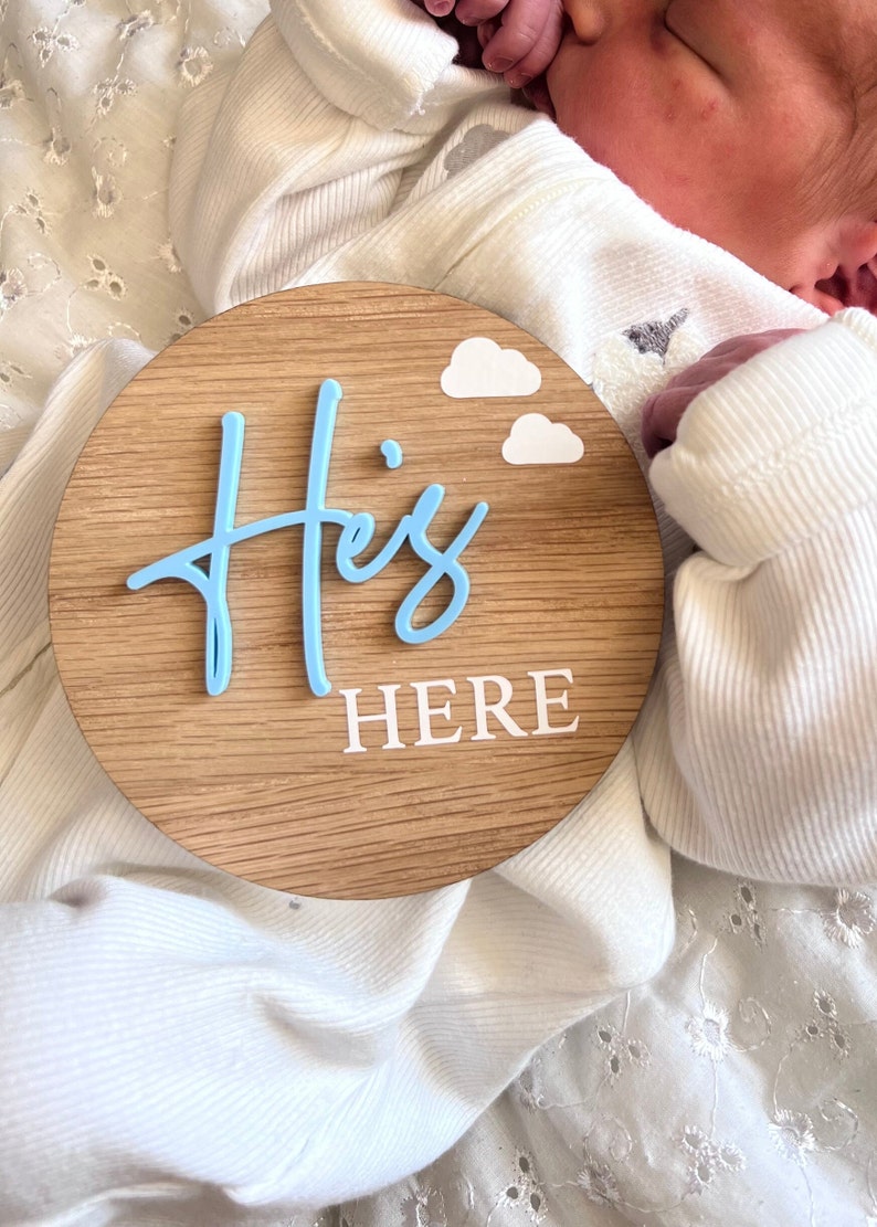 May include: A wooden round sign with the words "He's Here" in white lettering. The word "He's" is in light blue and is stylized. There are two white clouds on the sign.