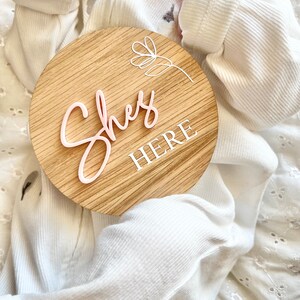 May include: A round wooden sign with the words "She's Here" in pink lettering. The sign has a white floral design and is being held by a baby wearing a white onesie.