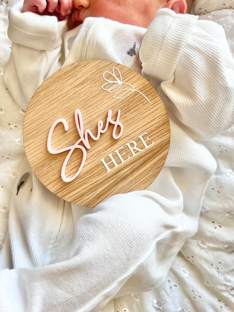 May include: A round wooden sign with the words "She's Here" in pink lettering. The sign has a white floral design and is being held by a baby wearing a white onesie.