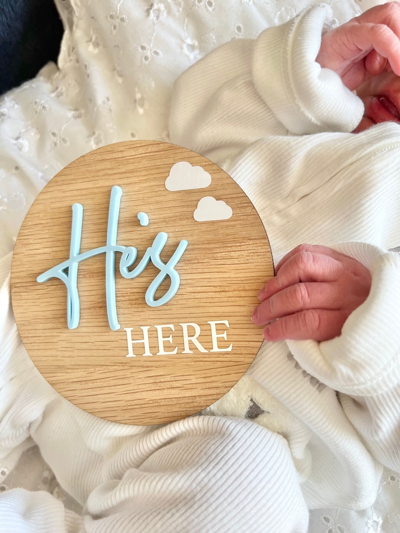 May include: A wooden round sign with the words "He's Here" in white lettering and two blue clouds. The sign is held by a baby's hand.
