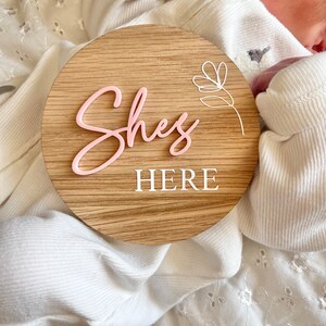 May include: A wooden round sign with the words "She's Here" in pink and white lettering. A white floral design is in the upper right corner of the sign.