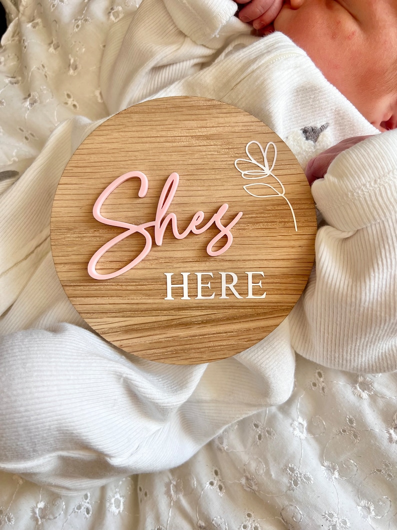 May include: A wooden round sign with the words "She's Here" in pink and white lettering. A white floral design is in the upper right corner of the sign.