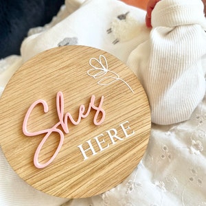 May include: A wooden round sign with the words "She's Here" in pink and white lettering. The sign has a simple floral design in white.