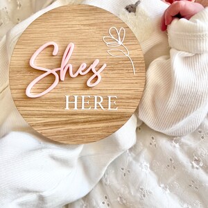 May include: A round wooden sign with the words "She's Here" in pink and white lettering. A white floral design is in the upper right corner of the sign.