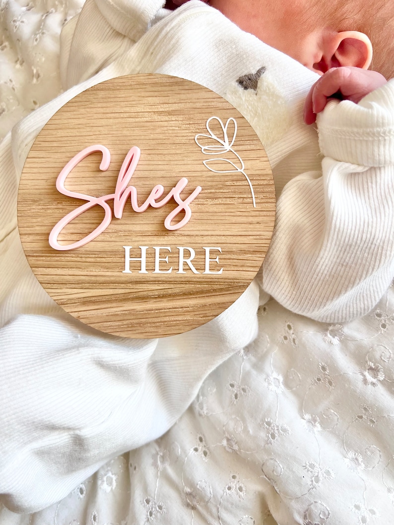 May include: A round wooden sign with the words "She's Here" in pink and white lettering. A white floral design is in the upper right corner of the sign.