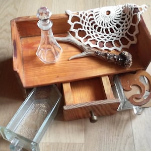 Op de afbeelding: Een houten kist met een open lade, een glazen fles met een kristallen stop, een wit kleedje en een geweistak. Een glazen bak en een houten decoratief element zijn ook aanwezig.