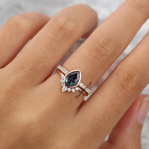 Pode incluir: Um conjunto de an&eacute;is em ouro rosa com uma pedra central em forma de pera de cor azul-esverdeada, rodeada por um halo de pequenas pedras transparentes de corte redondo. A banda &eacute; adornada com pedras adicionais ao longo do aro do anel.