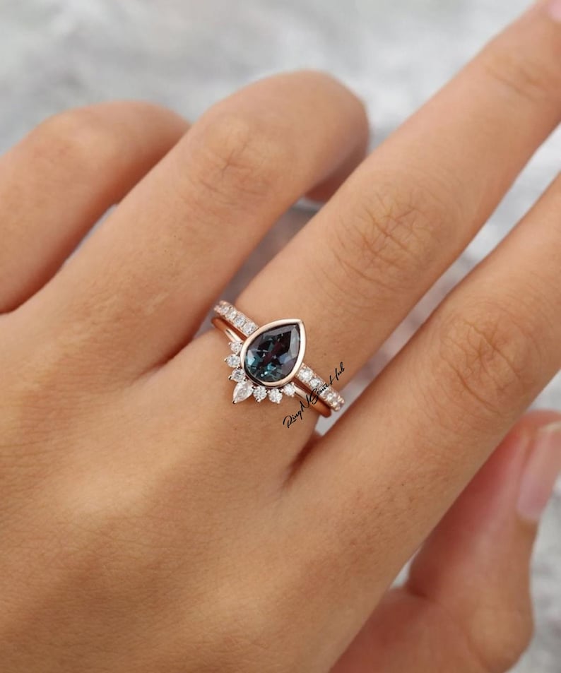 Pode incluir: Um conjunto de an&eacute;is em ouro rosa com uma pedra central em forma de pera de cor azul-esverdeada, rodeada por um halo de pequenas pedras transparentes de corte redondo. A banda &eacute; adornada com pedras adicionais ao longo do aro do anel.