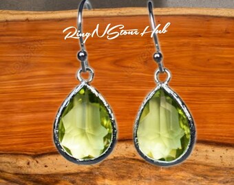 May include: A pair of silver teardrop earrings with green gemstone drops. The earrings are on a wooden surface.