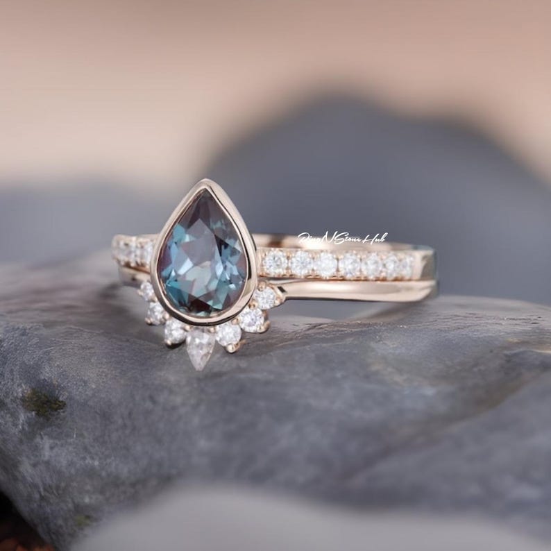 Pode incluir: Um conjunto de an&eacute;is em ouro rosa com uma pedra central azul-esverdeada em forma de pera numa crava&ccedil;&atilde;o de bisel, acentuada por um halo de pequenas pedras brancas redondas e uma alian&ccedil;a a condizer com uma fila de pedras brancas. Texto GemsHub vis&iacute;vel.