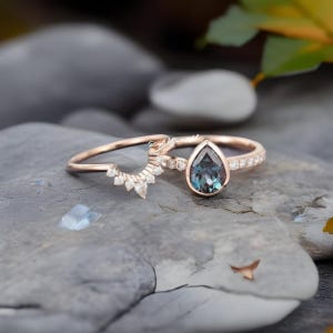 Pode incluir: Um conjunto de an&eacute;is em ouro rosa com uma pedra central azul-esverdeada em forma de pera rodeada por pequenas pedras claras na banda, acompanhado por uma banda curva a condizer adornada com v&aacute;rias pequenas pedras claras em leque.