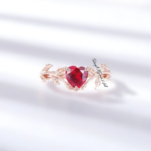 May include: A rose gold ring featuring a heart-shaped, deep red gemstone. The gemstone is set within a delicate, leaf-like design. The ring is displayed on a white background, highlighting its intricate details.