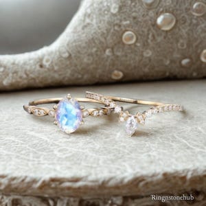 May include: A gold ring set featuring an oval moonstone center gem with a blue iridescent glow, flanked by small round diamonds on the band. A matching curved wedding band with marquise and round diamonds sits beside it, both marked Ringnstonehub.