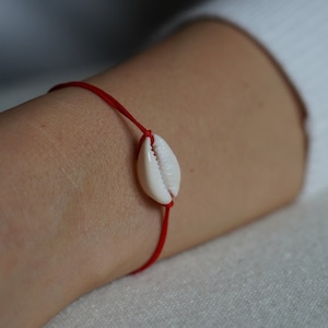 Könnte beinhalten: Ein rotes Armband mit einem weißen Muschel-Anhänger.