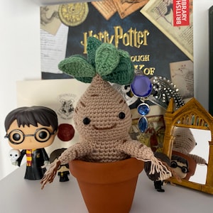 May include: A crocheted brown and green Mandrake plant in a terracotta pot. The plant has a smiling face and is sitting on a shelf with other Harry Potter themed items.