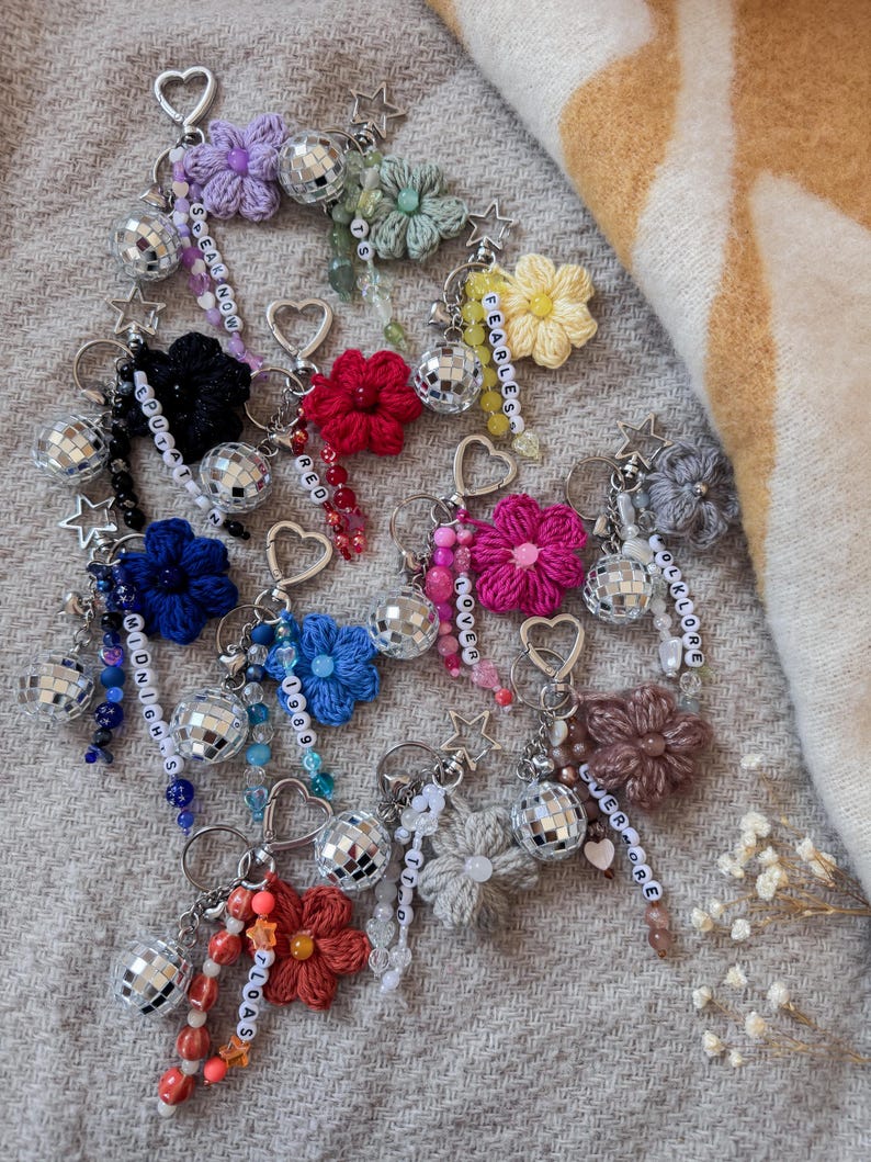 May include: A collection of handmade keychains featuring crocheted flowers in various colors, disco ball charms, and beaded accents. Each keychain has a heart-shaped clasp and a word or phrase spelled out in small beads.