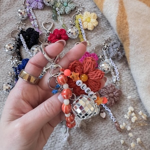 May include: A collection of handmade keychains featuring crocheted flowers, disco ball charms, and beaded accents. Some keychains include star and heart-shaped charms, with beaded text spelling out words like "FEARLESS" and "TLOAS".