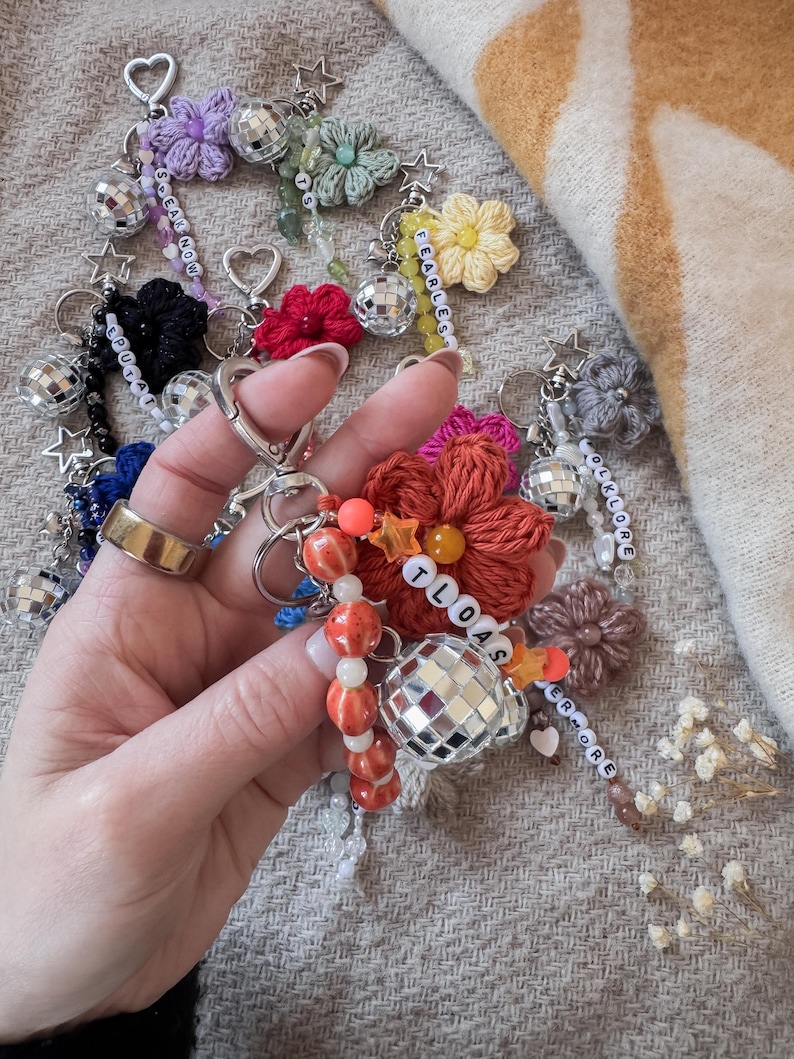 May include: A collection of handmade keychains featuring crocheted flowers, disco ball charms, and beaded accents. Some keychains include star and heart-shaped charms, with beaded text spelling out words like "FEARLESS" and "TLOAS".