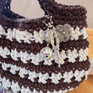 May include: A handmade crochet tote bag in brown and white stripes. The bag features a star-shaped charm, a disco ball, and a beaded word charm that says "FOLKLORE". The bag has a label that says "Made with love".