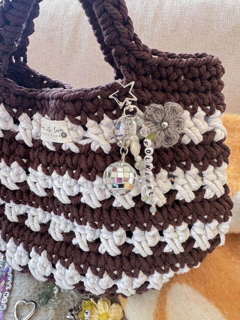 May include: A handmade crochet tote bag in brown and white stripes. The bag features a star-shaped charm, a disco ball, and a beaded word charm that says "FOLKLORE". The bag has a label that says "Made with love".