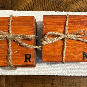 May include: Set of four wooden coasters with a natural wood grain finish. The coasters are tied together with twine and have the letters "R" and "M" burned into the wood.