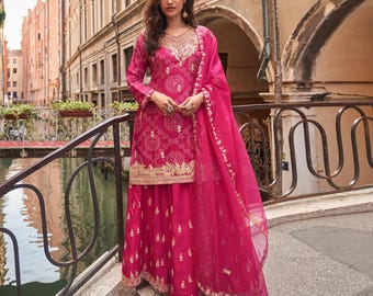 Può includere: Un completo indiano magenta ricamato con una dupatta abbinata. Il completo presenta intricati ricami dorati sullo scollo, sulle maniche e sull'orlo. La modella è in piedi su un ponte di fronte a un canale.