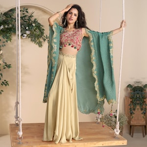 Puede incluir: Conjunto femenino con un top corto rosa y dorado bordado, una falda beige fluida y una capa turquesa con ribetes dorados. El atuendo se exhibe en un columpio de madera, mostrando una mezcla de texturas y colores.