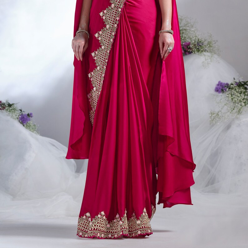 May include: A vibrant fuchsia-colored saree with intricate gold embroidery along the borders and a decorative panel. The saree features a draped design with a flowing cape-like element. The image showcases a close-up view of the garment.