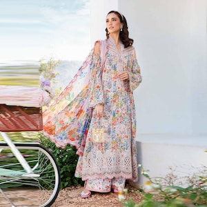 Puede incluir: Una mujer lleva un vestido de estampado floral blanco y rosa con una bufanda de estampado floral transparente. El vestido tiene un borde de encaje y la mujer lleva una pequeña caja blanca y dorada.