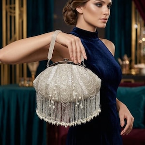 Può includere: Una borsa da sera argentata con perline e frange di perle. La borsa ha un manico argentato e una chiusura a scatto. La borsa è tenuta da una persona che indossa un abito di velluto blu scuro.