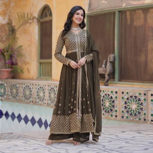 Puede incluir: Un traje Anarkali bordado en verde oliva con detalles dorados. El vestido de manga larga presenta intrincados bordados dorados en el corpiño y el dobladillo, combinado con pantalones palazzo a juego y una dupatta. El conjunto es adecuado para ocasiones festivas.