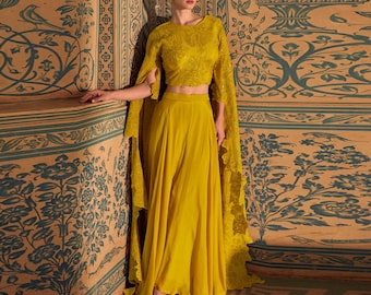 May include: A yellow crop top with a cape-like overlay and a matching yellow skirt. The top is embroidered with a floral design and the skirt is flowing and pleated. The model is standing in front of a patterned wall.