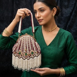 May include: A decorative burgundy velvet handbag with a pearl-beaded handle and fringe. The bag is adorned with gold and emerald green embellishments. The background is a dark, textured surface.