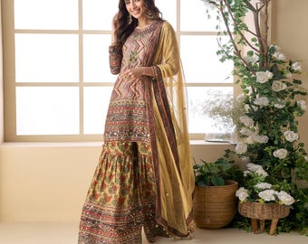 May include: A woman wearing a traditional Indian salwar kameez. The outfit features a long-sleeved tunic with intricate embroidery and a matching palazzo pant. The ensemble is in shades of beige, red, and gold, with a sheer dupatta.