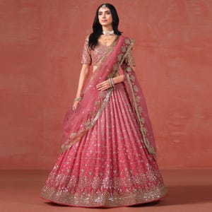 Op de afbeelding: Een roze en gouden lehenga choli, een traditionele Indiase outfit. De rok heeft een roze gradiënt, versierd met ingewikkelde gouden borduursels en spiegelwerk. De bijpassende dupatta en blouse hebben vergelijkbare details.