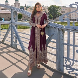 Puede incluir: Un vestido largo color ciruela con bordados y adornos dorados. El vestido presenta un escote en V, mangas largas y una falda fluida. El bordado dorado es intrincado y detallado, y el vestido se combina con sandalias doradas y una bufanda a juego.