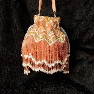 Puede incluir: Un bolso de mano con cuentas de color melocotón con cierre de cordón y flecos decorativos. El bolso presenta detalles de lentejuelas doradas y blancas y un ribete blanco con forma de perla en la parte inferior. El bolso tiene una correa de color melocotón.