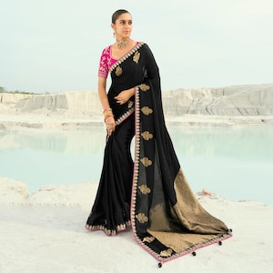 May include: A black saree with gold embroidery and a pink border. The saree features a pink blouse with gold embroidery on the sleeves. The garment is draped over a person standing in front of a body of water.
